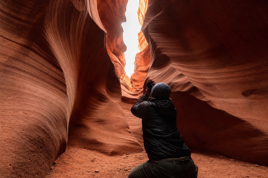 Visitor crouching low to photograph upward through curving canyon walls toward the sky