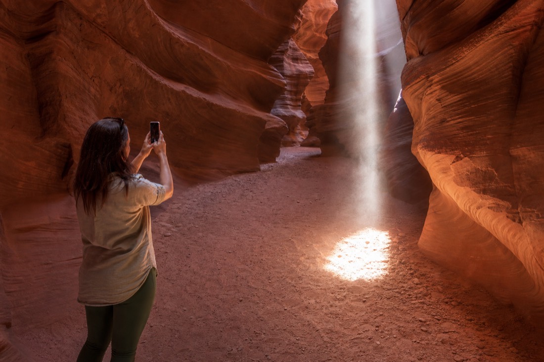 Visitor photographing a sunbeam in a slot canyon using only a handheld phone