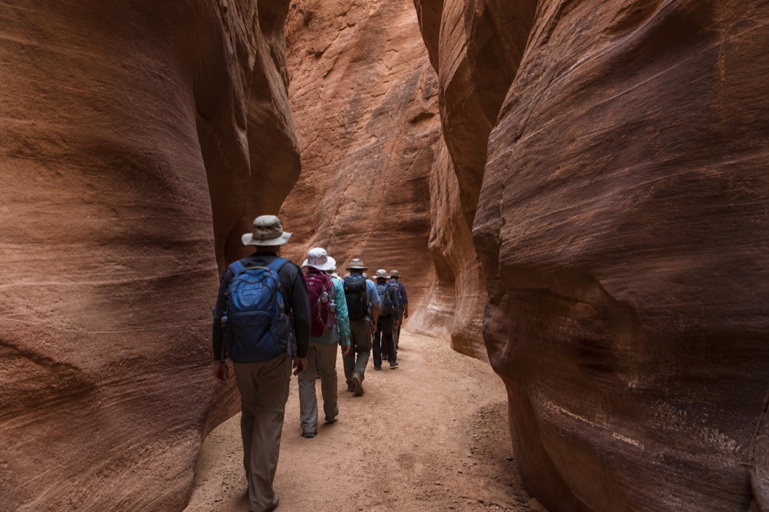 Small tour group walking single file along the sandy floor of a narrow slot canyon