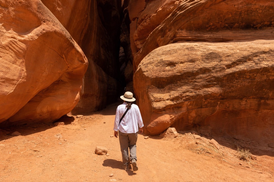 Visitor in light layers and a sun hat walking toward a slot canyon entrance in bright desert light