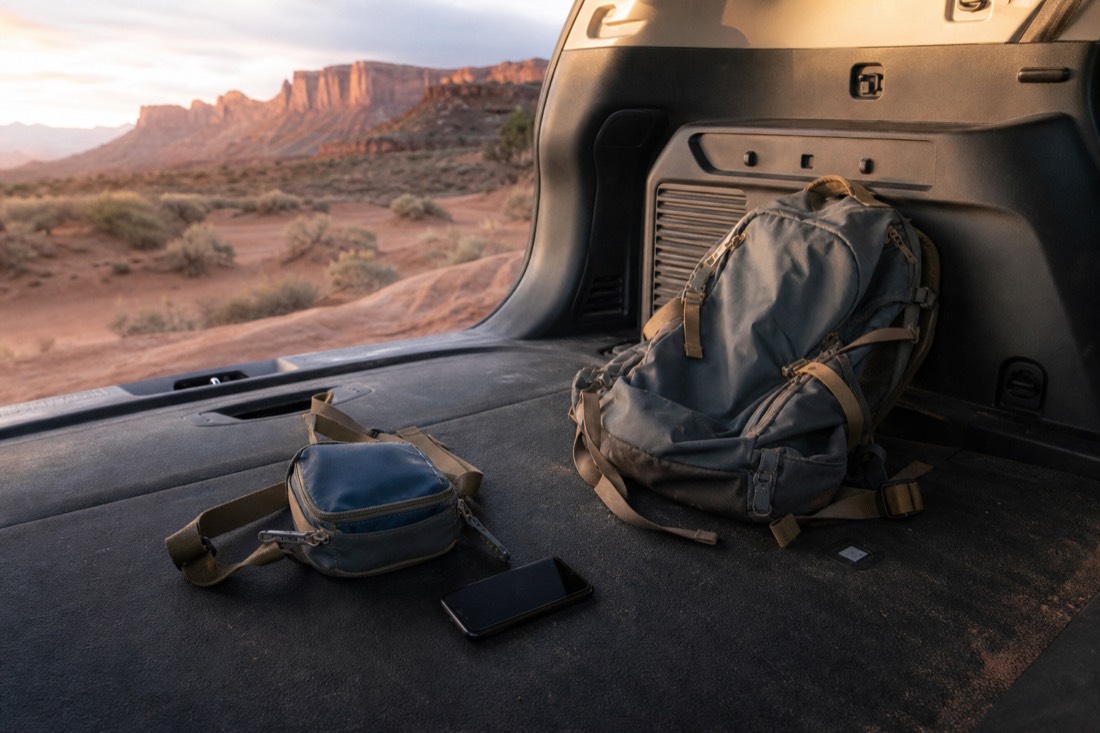 Small sling bag and phone laid out next to a large backpack left behind in a car trunk