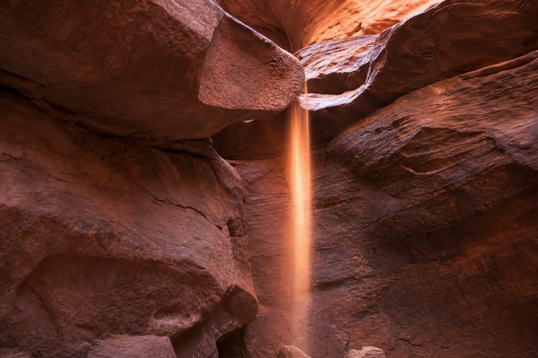 Fine red sand streaming down a sandstone canyon wall in a thin ribbon of light