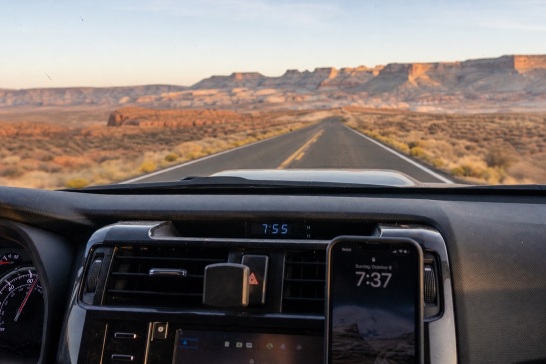 Car dashboard clock and phone showing different times on a desert highway near Page