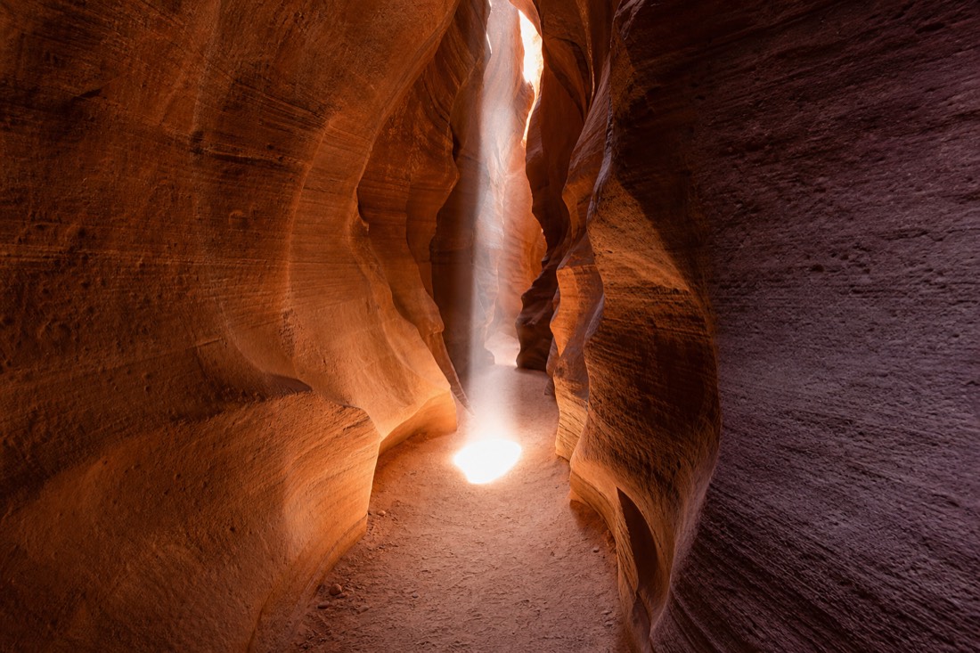 Shaft of sunlight reaching the sandy floor of a narrow sandstone slot canyon at midday