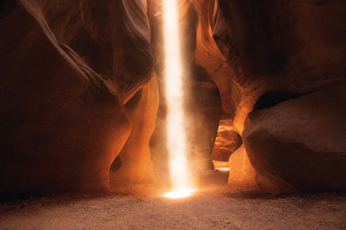 Bright vertical sunbeam reaching the floor of a slot canyon amid swirling illuminated dust