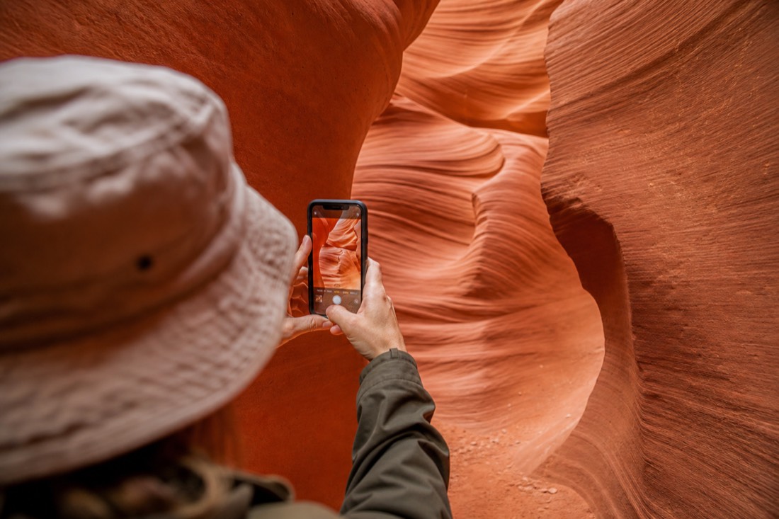 Visitor framing a swirl of orange sandstone on a phone screen inside a narrow slot canyon