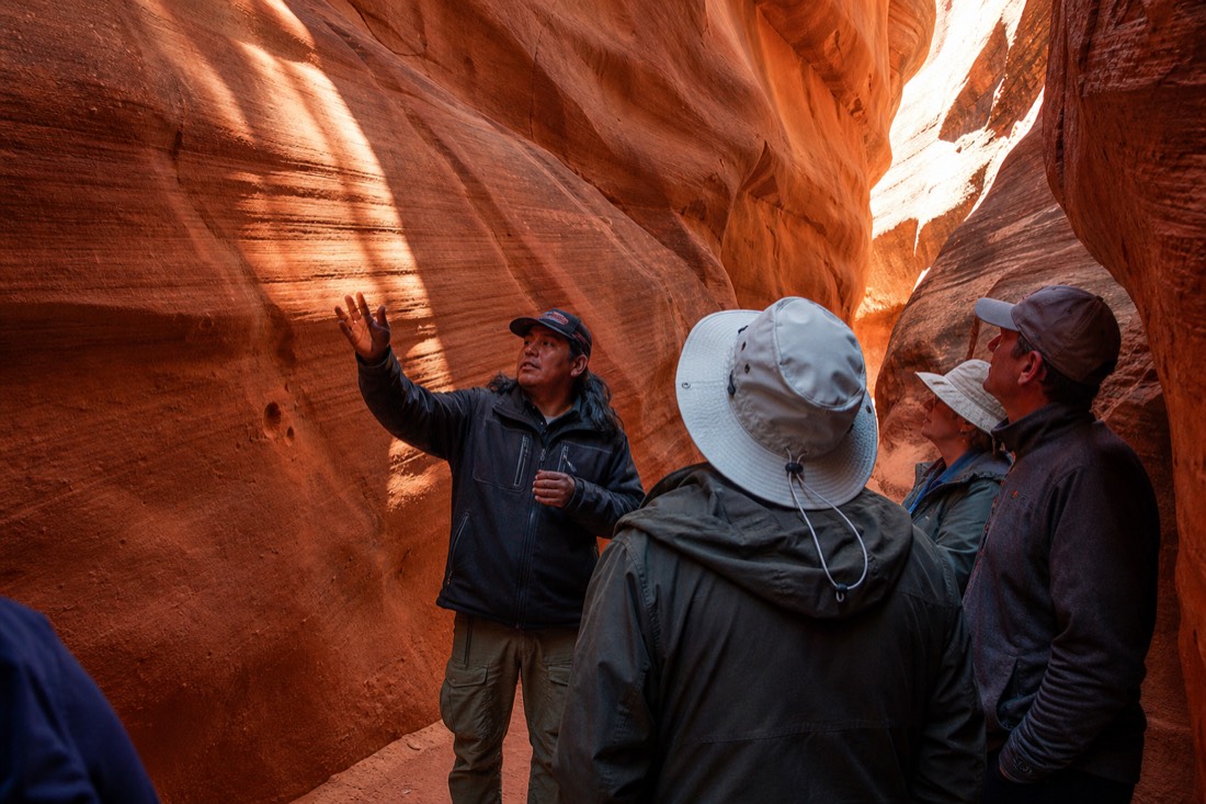 Navajo guide gesturing toward canyon light patterns for a small tour group inside a slot canyon