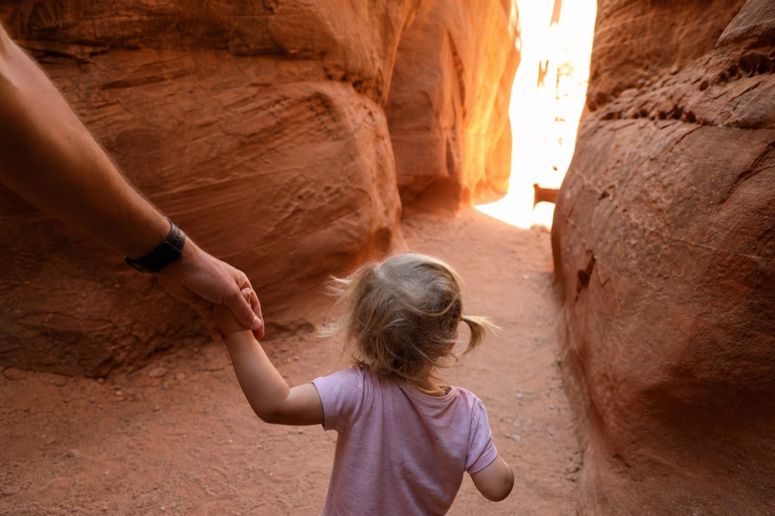 Young child holding a parent's hand at the sunlit entrance of a sandstone slot canyon