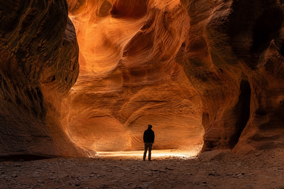 Lone visitor silhouetted in a quiet slot canyon chamber with layered orange rock walls