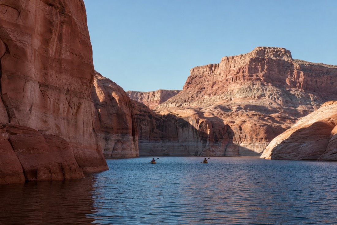 Kayakers paddling calm blue water between tall red sandstone canyon walls on Lake Powell